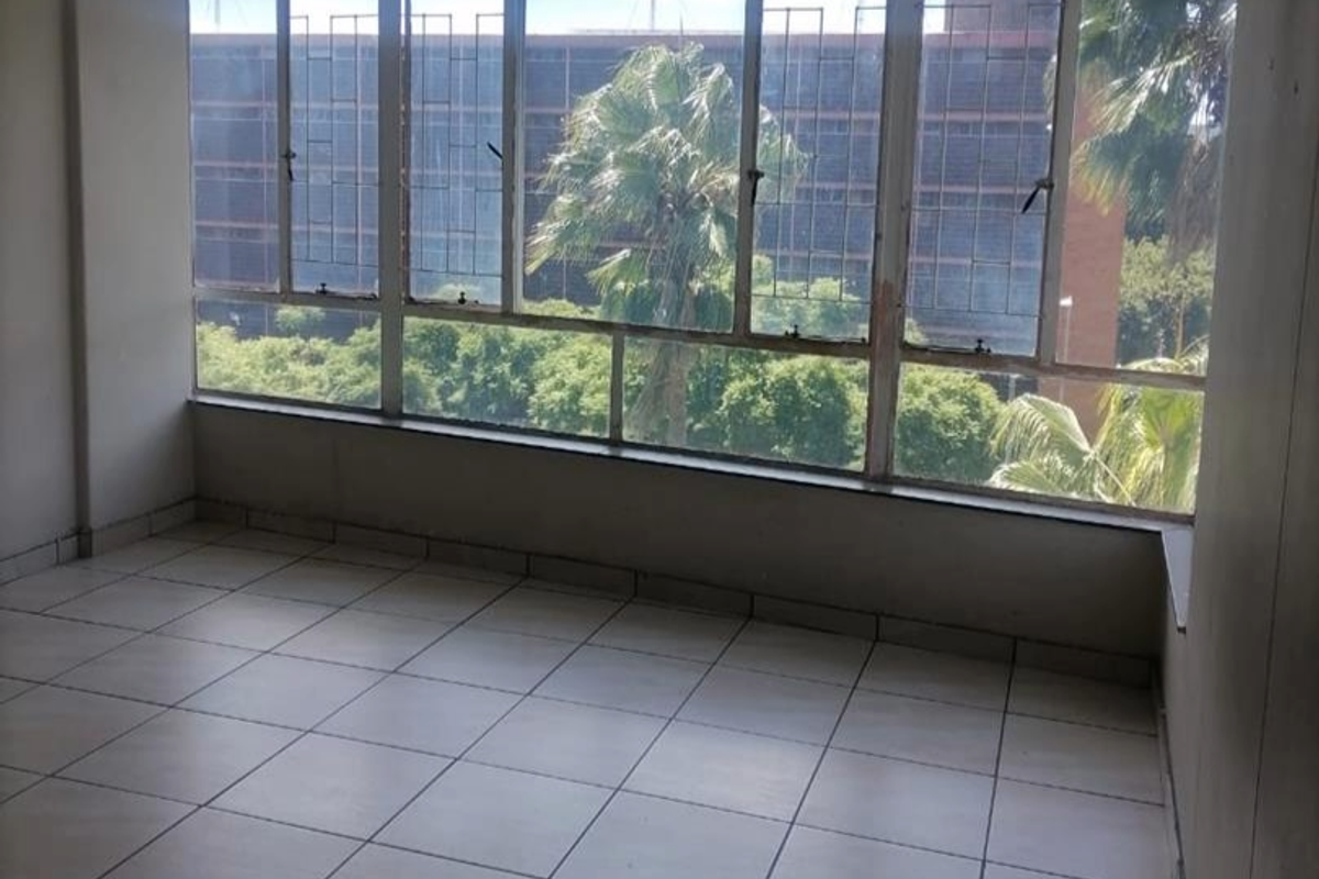An empty, sunlit room featuring large metal-framed windows and tiled flooring, offering a view of the surrounding urban landscape.
