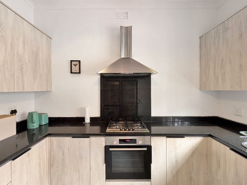 A modern kitchen featuring light wood-grain cabinetry, black granite countertops, a built-in oven, gas hob, and a stainless steel extractor fan.