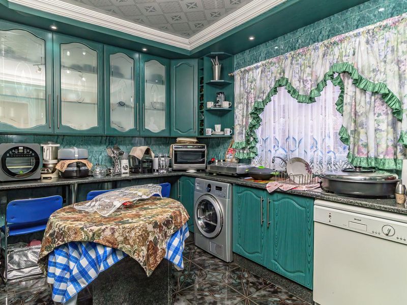 A dated kitchen featuring teal cabinetry, granite countertops, integrated appliances, and decorative ceiling tiles, offering a functional layout with significant potential for modernization.