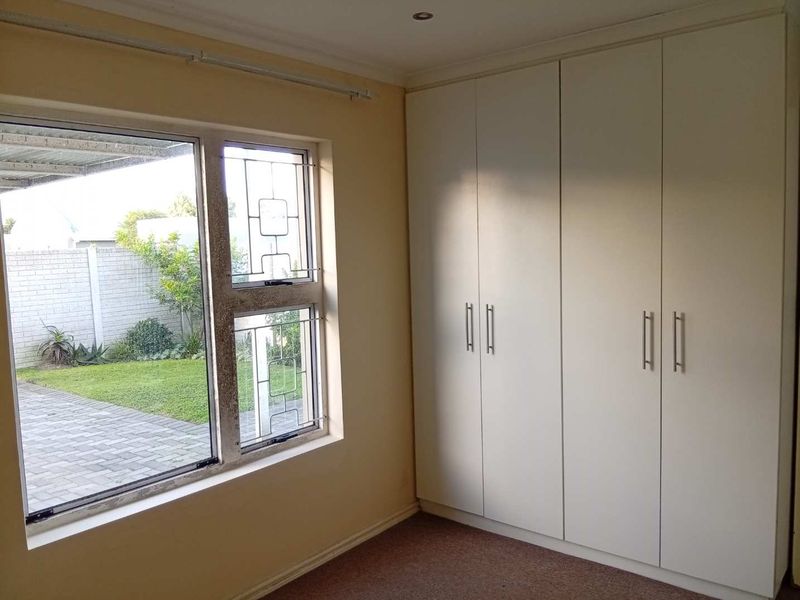 A bright, empty bedroom featuring built-in cupboards, carpeted flooring, and large windows with security bars overlooking the garden area.