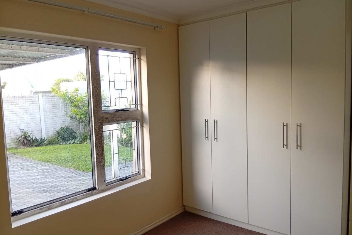 A bright, empty bedroom featuring built-in cupboards, carpeted flooring, and large windows with security bars overlooking the garden area.