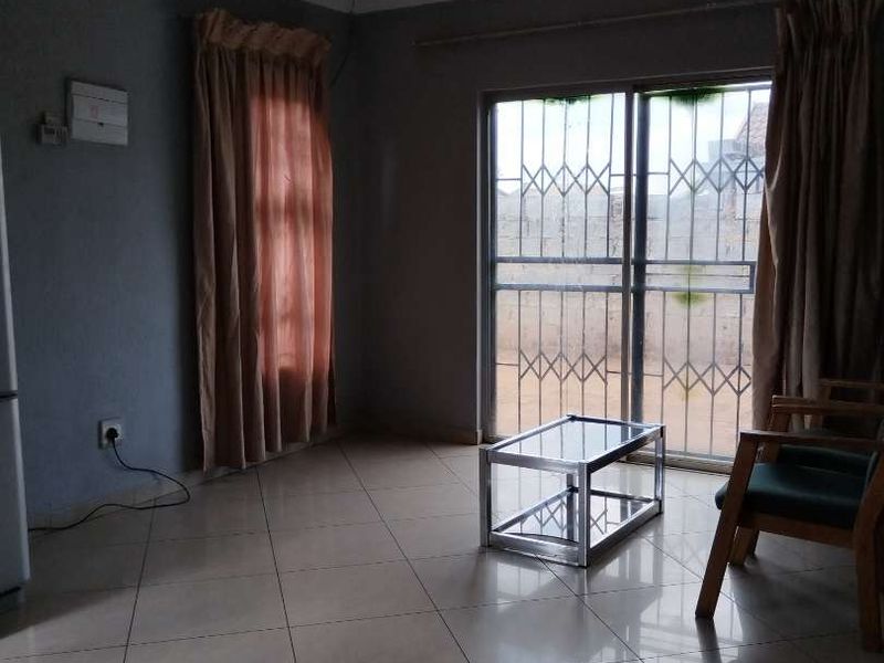 A sparsely furnished living room featuring tiled flooring and a sliding glass door with integrated security gate, providing access to the exterior.