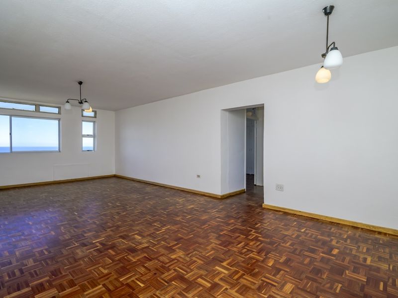 Spacious, empty living area featuring classic parquet flooring, neutral white walls, and large windows offering natural light and ocean views.