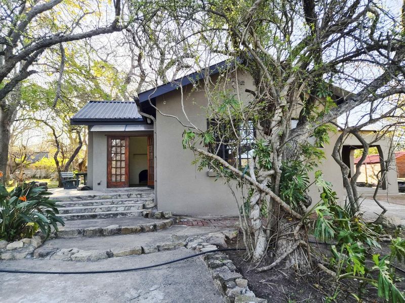 Rear view of the house featuring a stone-stepped entrance, timber-framed glass doors, and a mature garden setting with a concrete patio area.