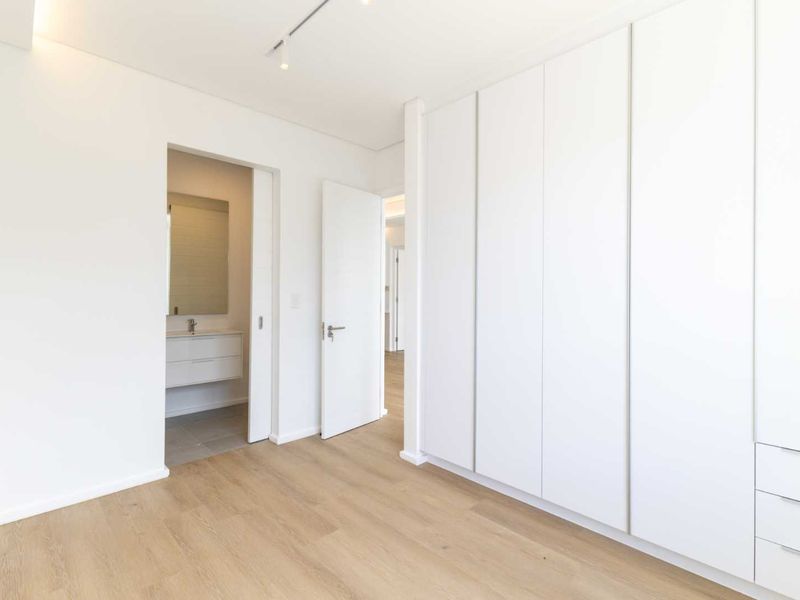 A bright, modern bedroom featuring extensive built-in white cabinetry, light wood-look laminate flooring, and direct access to an en-suite bathroom.