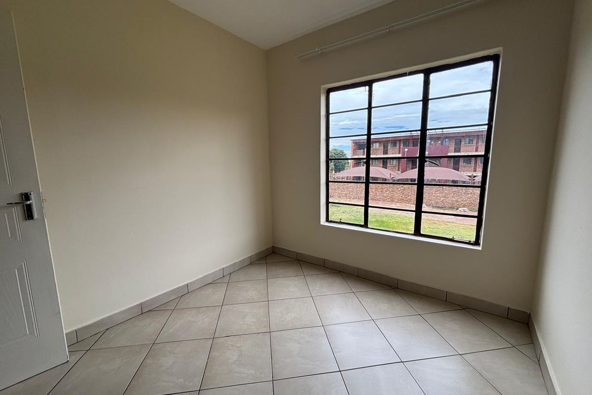 A bright, empty bedroom featuring neutral tiled flooring, a large window with metal frame, and a standard interior door.