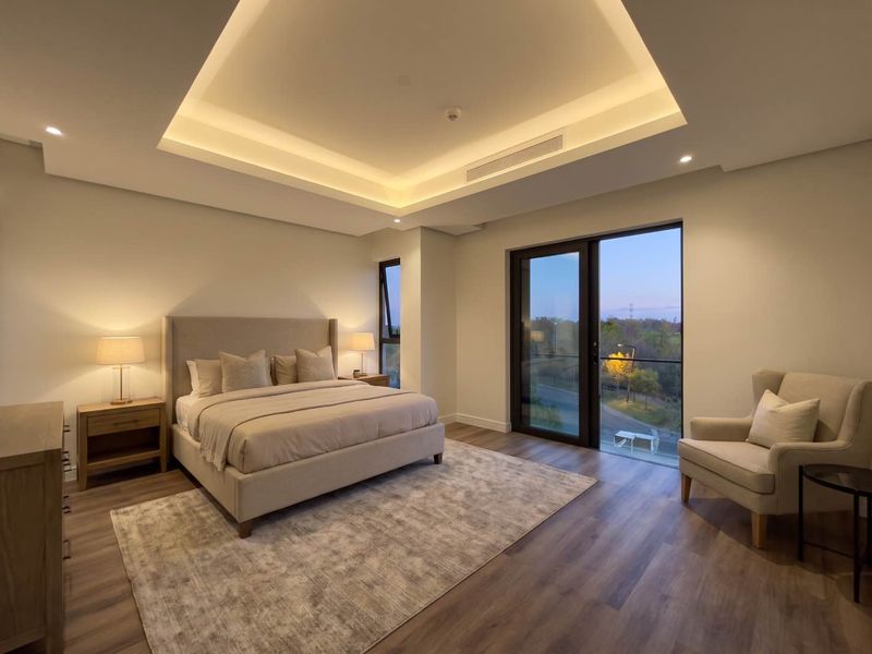Spacious modern bedroom featuring a recessed tray ceiling with ambient cove lighting, wood-look flooring, and large glass doors opening to a private balcony.