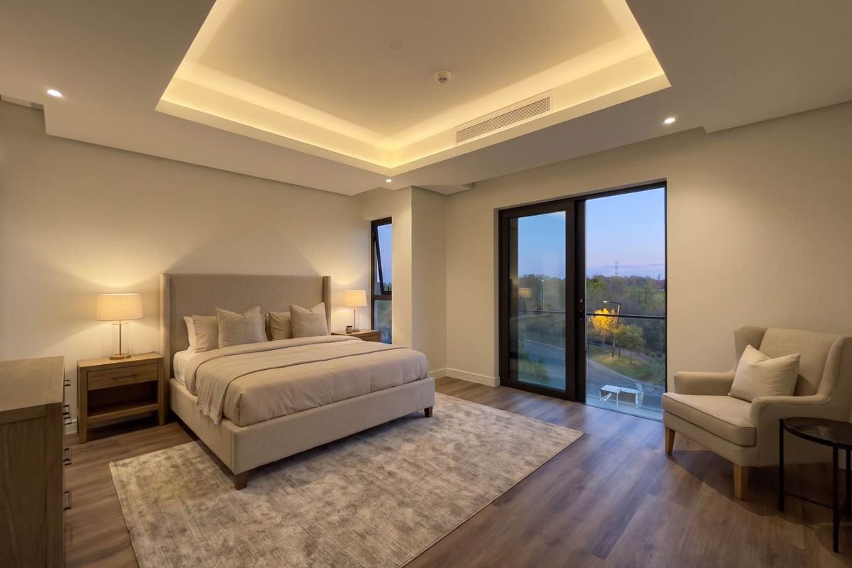 Spacious modern bedroom featuring a recessed tray ceiling with ambient cove lighting, wood-look flooring, and large glass doors opening to a private balcony.
