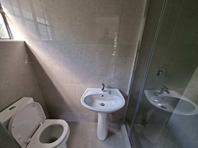A clean, modern bathroom featuring a pedestal basin, toilet, and a glass-enclosed shower with neutral wall and floor tiling.