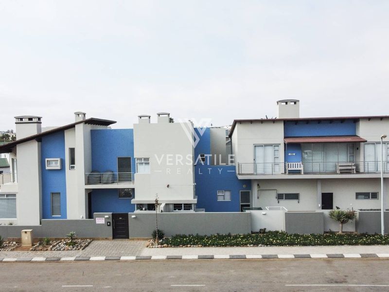 Front elevation of a modern two-story residential building featuring blue and grey facade, balconies with metal railings, and a secure boundary wall.