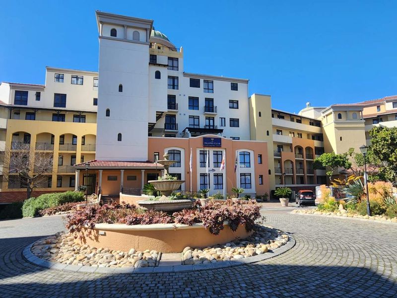 Front exterior view of the Island Club apartment complex featuring a paved driveway, central fountain, and multi-story building facade with balconies.