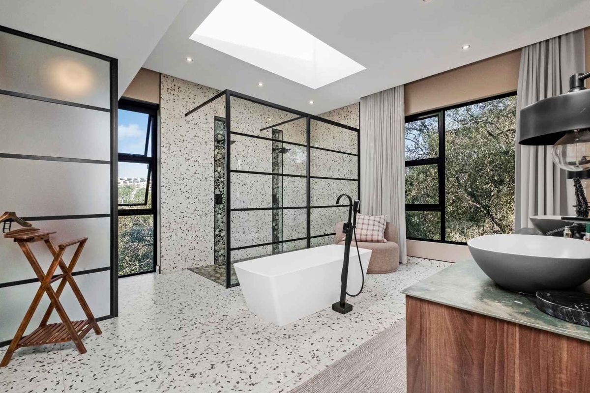 Modern luxury bathroom featuring a freestanding bathtub, walk-in shower with black frame, terrazzo-style flooring, and a large skylight providing natural light.