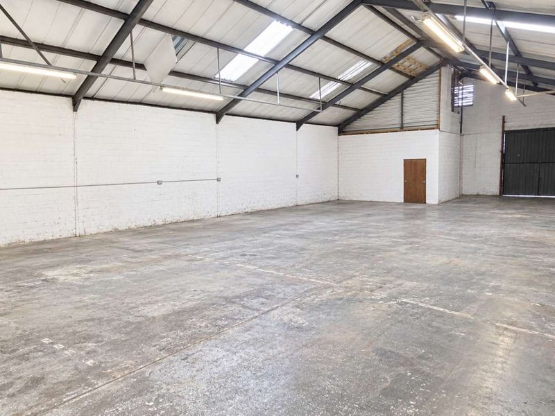 Spacious open-plan office or industrial workspace featuring exposed steel trusses, skylights, concrete flooring, and a large roller shutter door.
