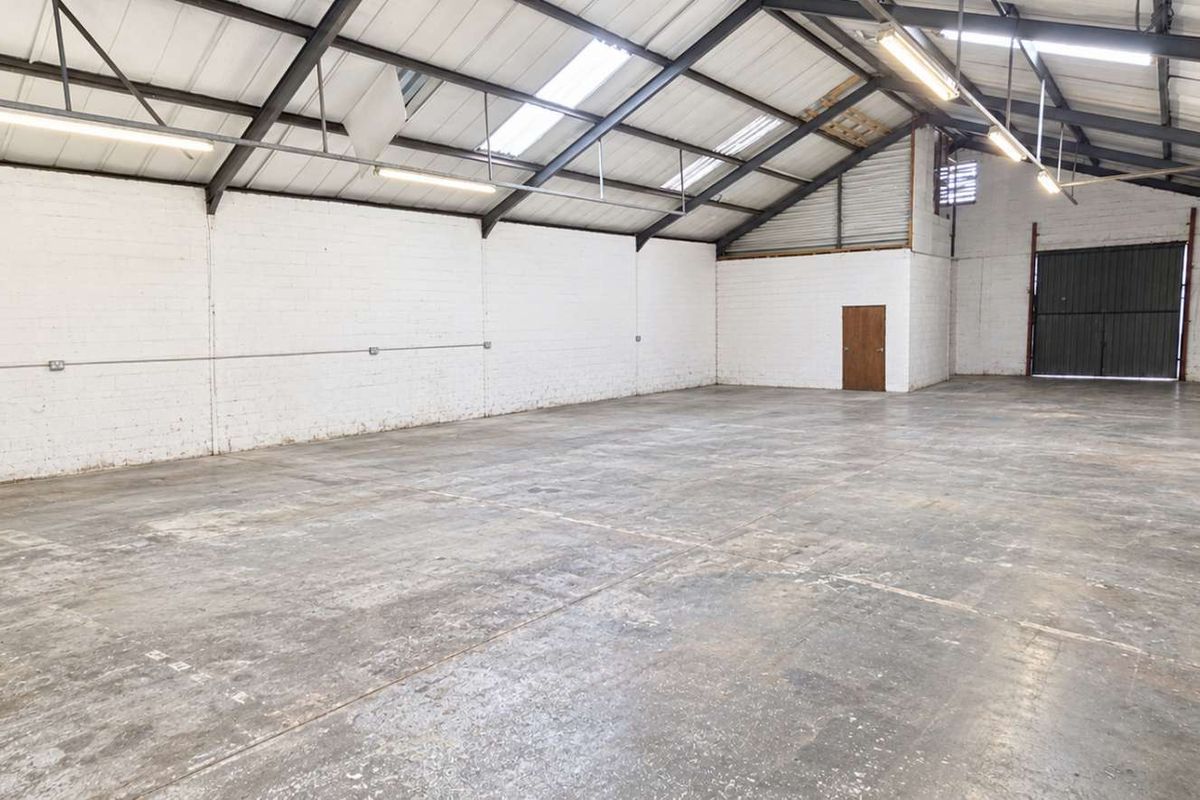Spacious open-plan office or industrial workspace featuring exposed steel trusses, skylights, concrete flooring, and a large roller shutter door.