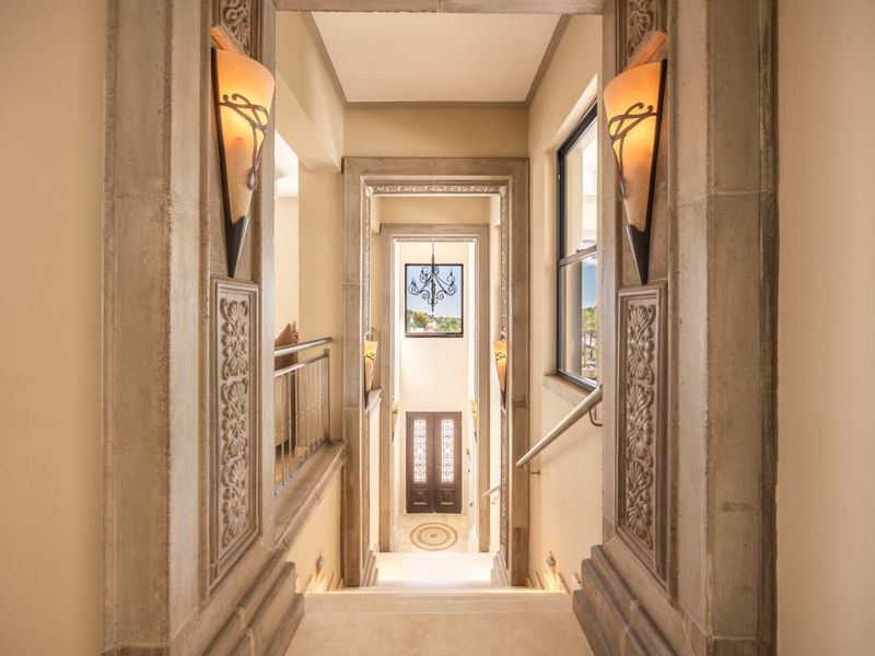 Elegant hallway featuring intricate stone archways, decorative wall carvings, and sophisticated wall sconces leading down a staircase with a metal handrail.
