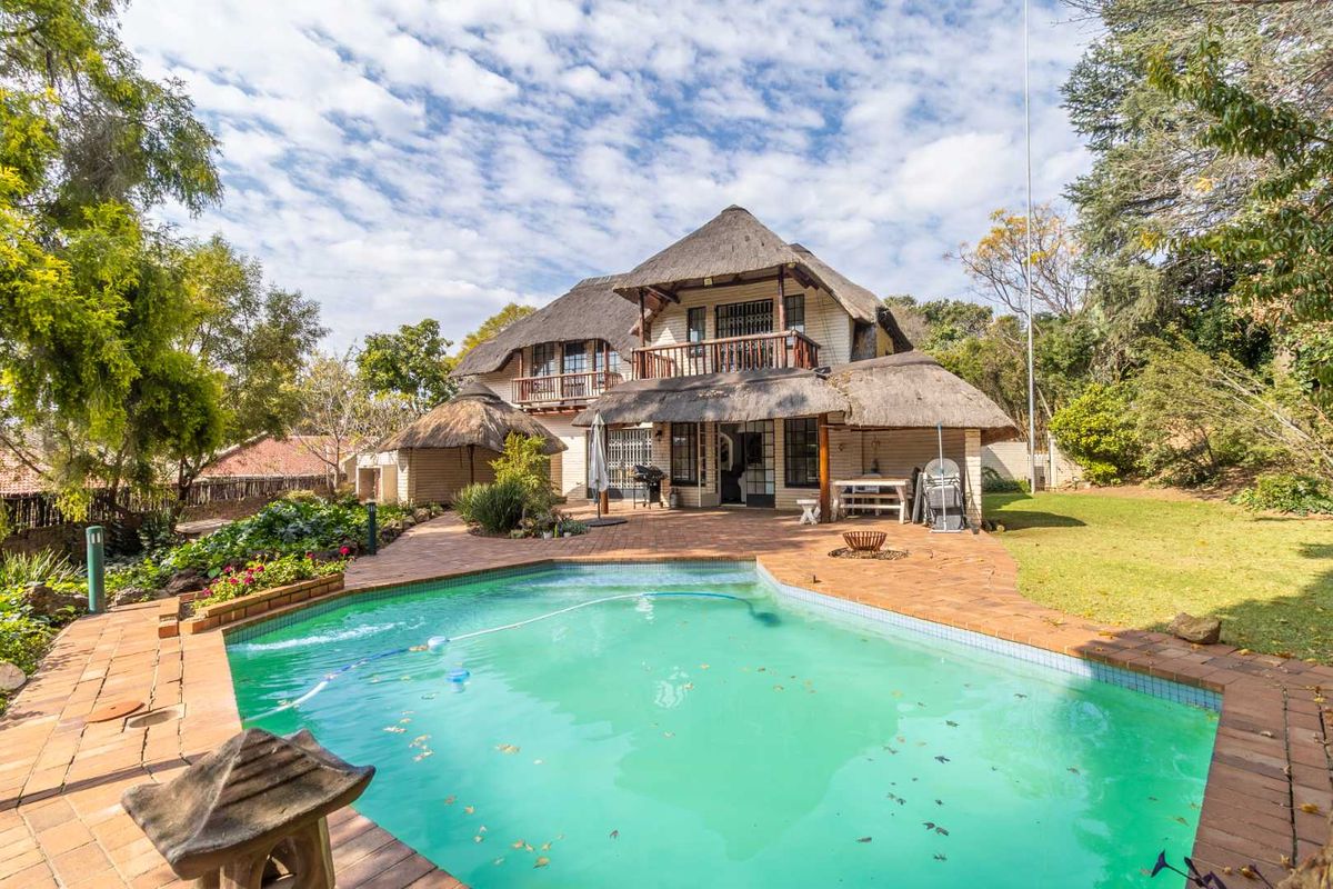 Rear view of a multi-story house featuring a thatched roof, a paved patio area, and a swimming pool set within a landscaped garden.