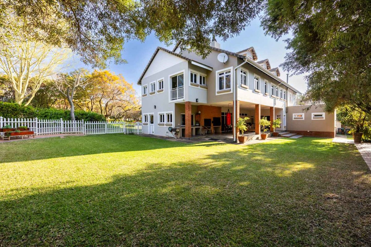 Rear view of a multi-story house featuring a spacious lawn, covered patio area with brick pillars, and a white picket fence.