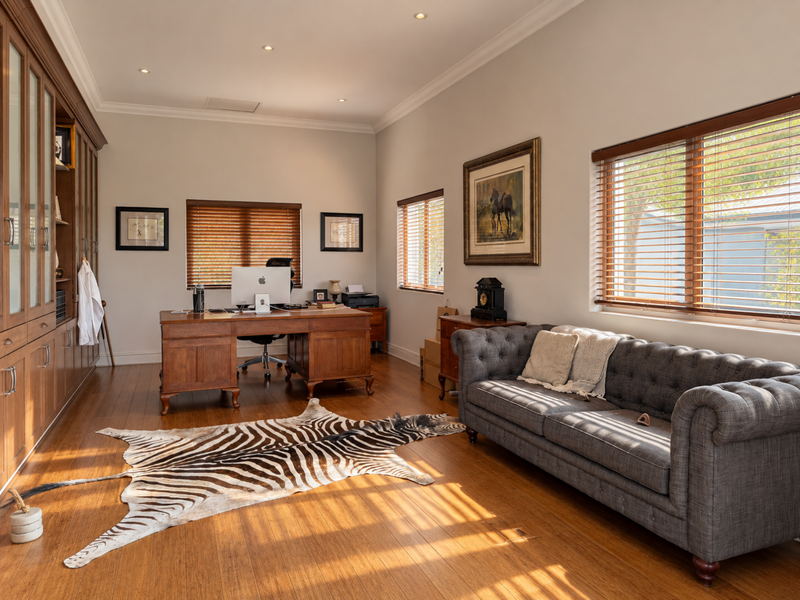 A well-appointed home office featuring extensive built-in cabinetry, warm hardwood flooring, recessed lighting, and large windows with venetian blinds.
