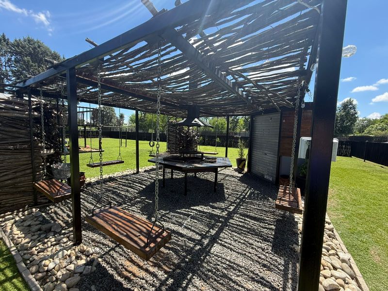 Outdoor entertainment area featuring a metal pergola with a rustic branch roof, built-in braai, and swing seating set over a gravel patio.