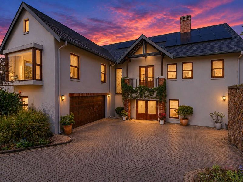 A well-maintained double-story residence featuring a paved driveway, integrated double garage, solar panels, and a charming brick and plaster facade at sunset.