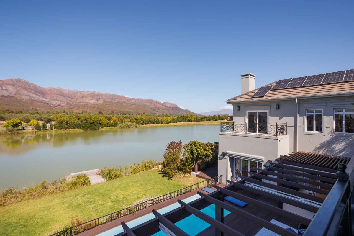 Rear view of the property featuring a swimming pool, timber pergola, balcony, and solar panels, overlooking a scenic lake and mountain landscape.