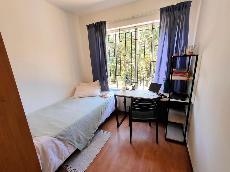 A compact bedroom featuring laminate flooring and a large window fitted with security bars, providing natural light and a view of the outdoors.