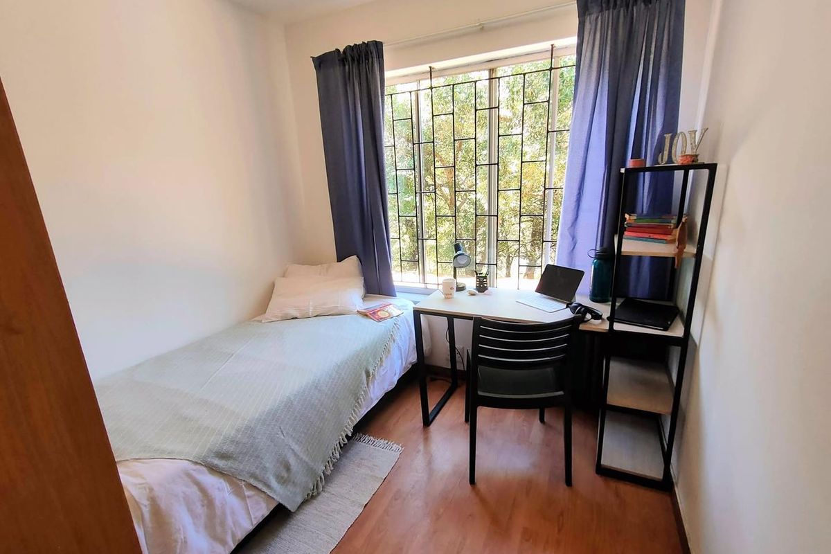 A compact bedroom featuring laminate flooring and a large window fitted with security bars, providing natural light and a view of the outdoors.