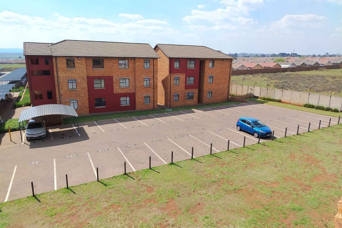 Aerial view of a multi-story brick apartment complex featuring a paved parking lot, carports, and perimeter fencing in a suburban setting.