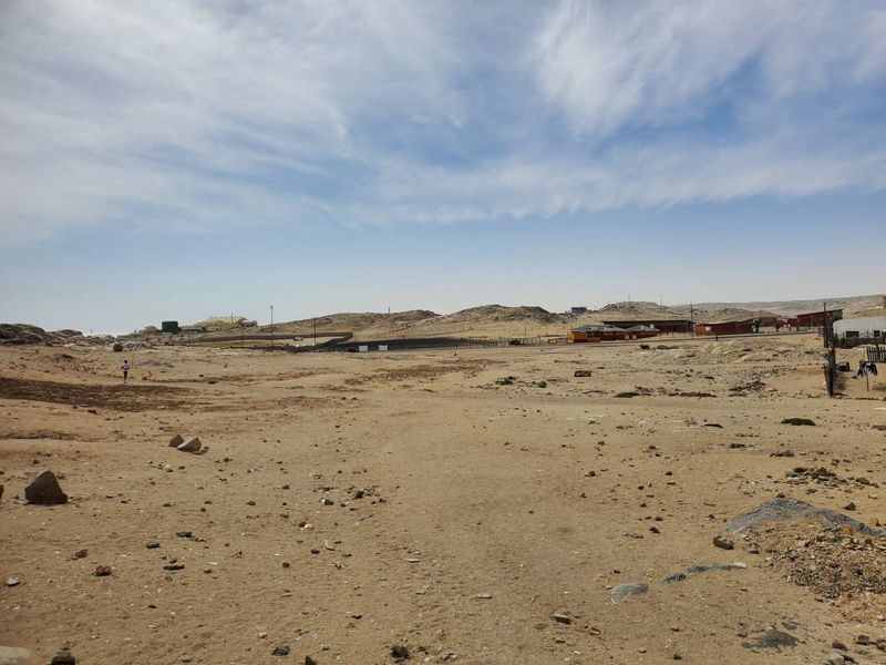 A wide-angle view of a vacant commercial land plot featuring natural terrain, perimeter fencing, and distant surrounding structures.