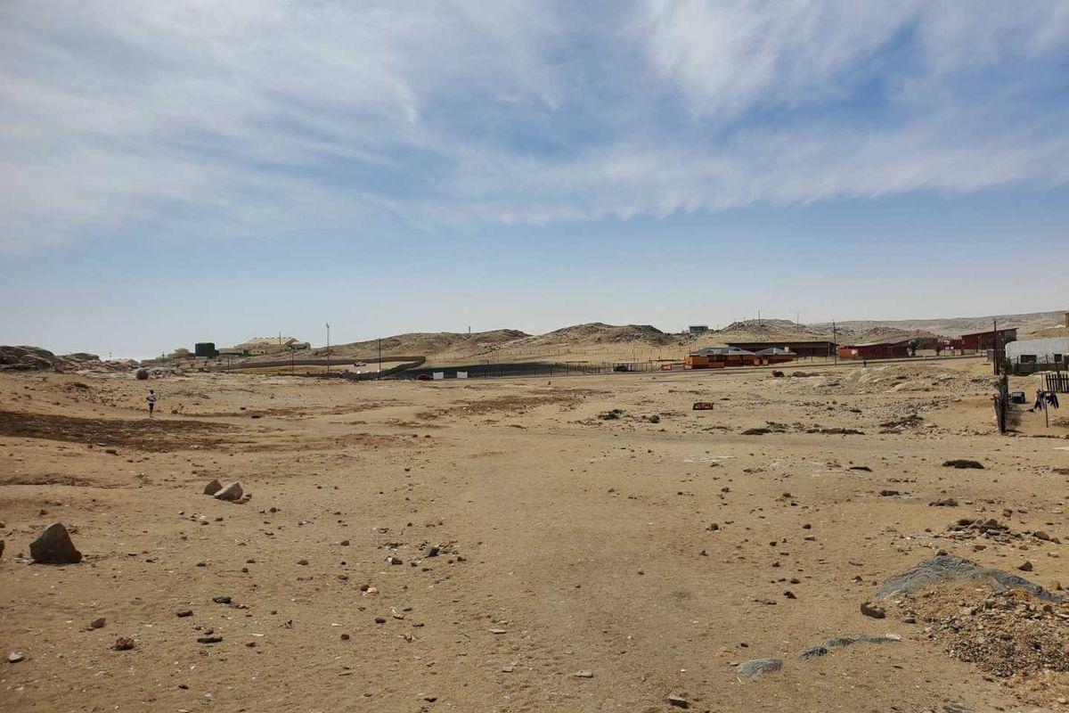 A wide-angle view of a vacant commercial land plot featuring natural terrain, perimeter fencing, and distant surrounding structures.
