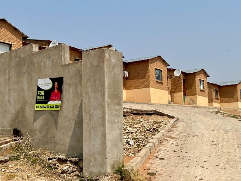 Street view of a townhouse complex featuring brick facades, corrugated metal roofing, and a paved driveway with an unfinished concrete boundary wall.