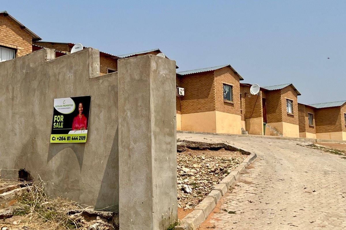 Street view of a townhouse complex featuring brick facades, corrugated metal roofing, and a paved driveway with an unfinished concrete boundary wall.
