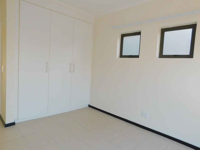 A clean, empty bedroom featuring neutral tiled flooring, built-in white cupboards, and two square windows with dark frames.