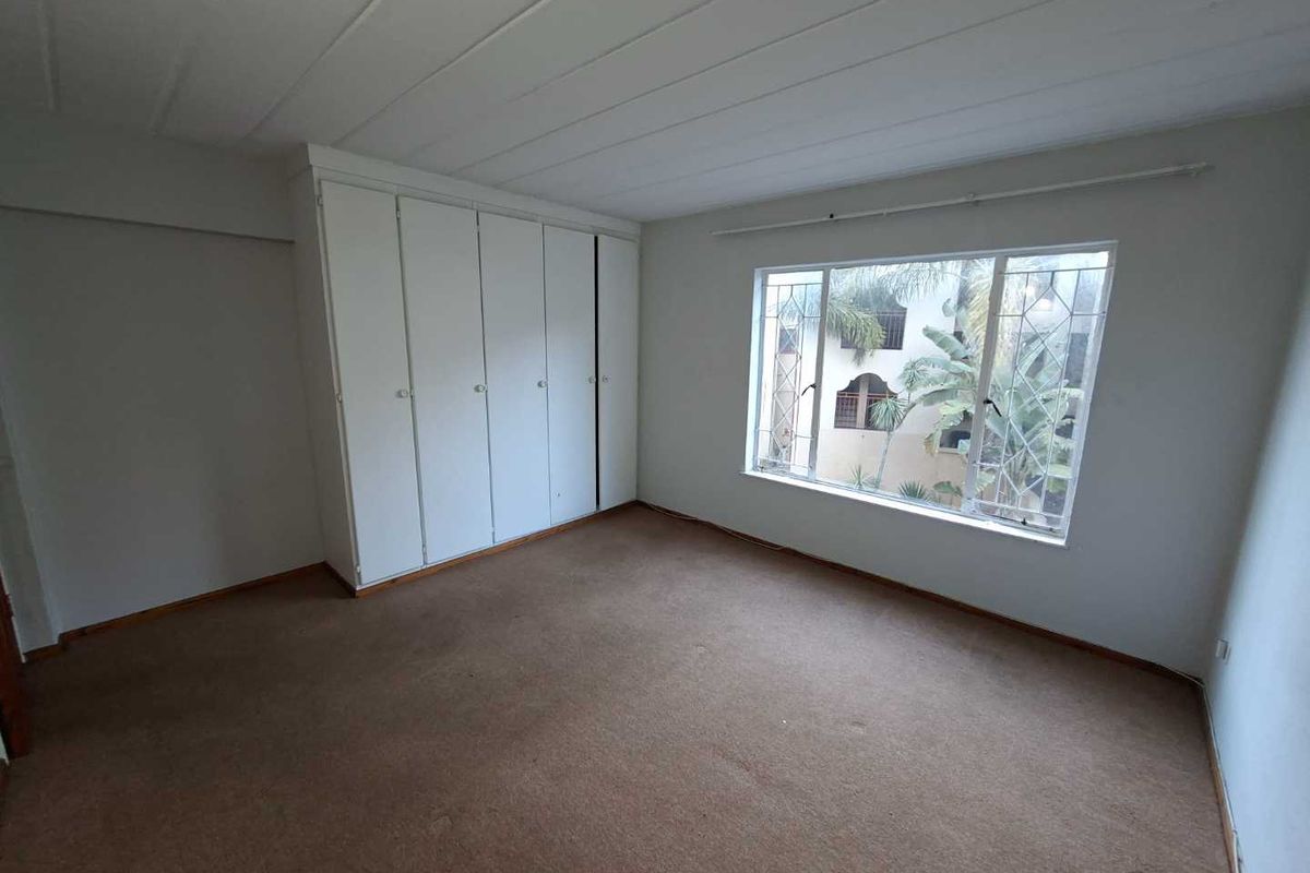 A spacious, empty bedroom featuring built-in cupboards, carpeted flooring, and a large window with security bars overlooking the exterior.