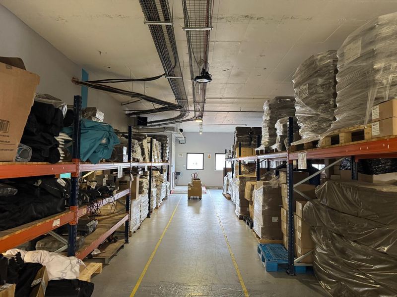 Industrial warehouse space featuring high ceilings with exposed cable trays, concrete flooring with safety markings, and extensive heavy-duty metal racking systems.