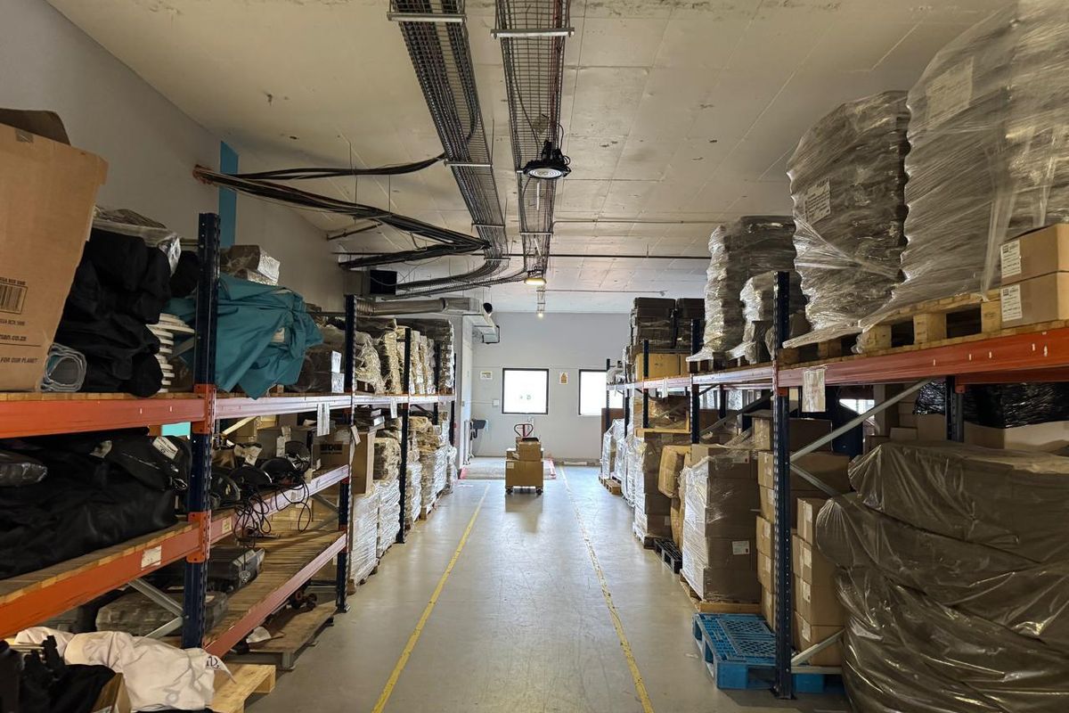 Industrial warehouse space featuring high ceilings with exposed cable trays, concrete flooring with safety markings, and extensive heavy-duty metal racking systems.