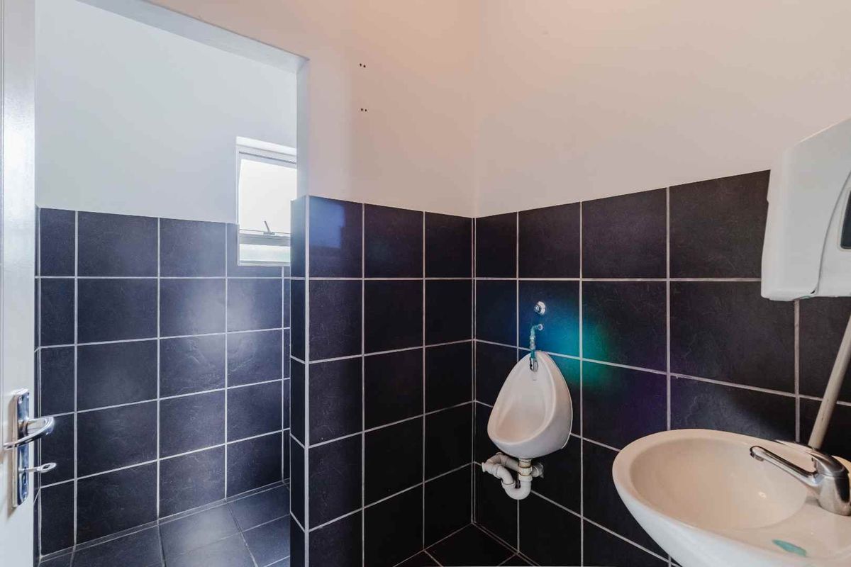 A functional commercial restroom featuring dark tiled walls, a wall-mounted urinal, and a small basin with chrome fixtures.