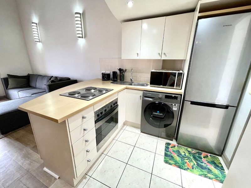 Compact open-plan kitchen featuring light-toned cabinetry, integrated electric hob and oven, stainless steel sink, and tiled flooring, seamlessly connected to the living area.