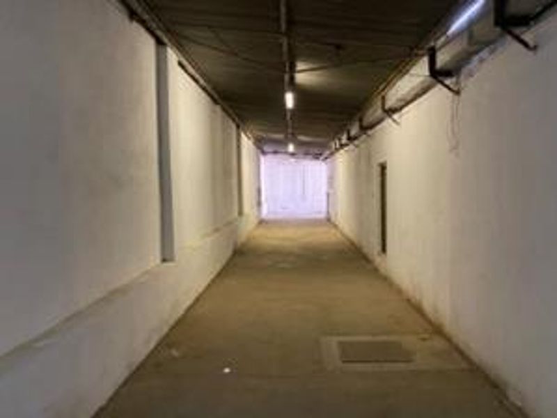 A long, industrial-style corridor featuring exposed ceiling infrastructure, concrete flooring, and basic fluorescent lighting, suitable for various commercial or storage access purposes.