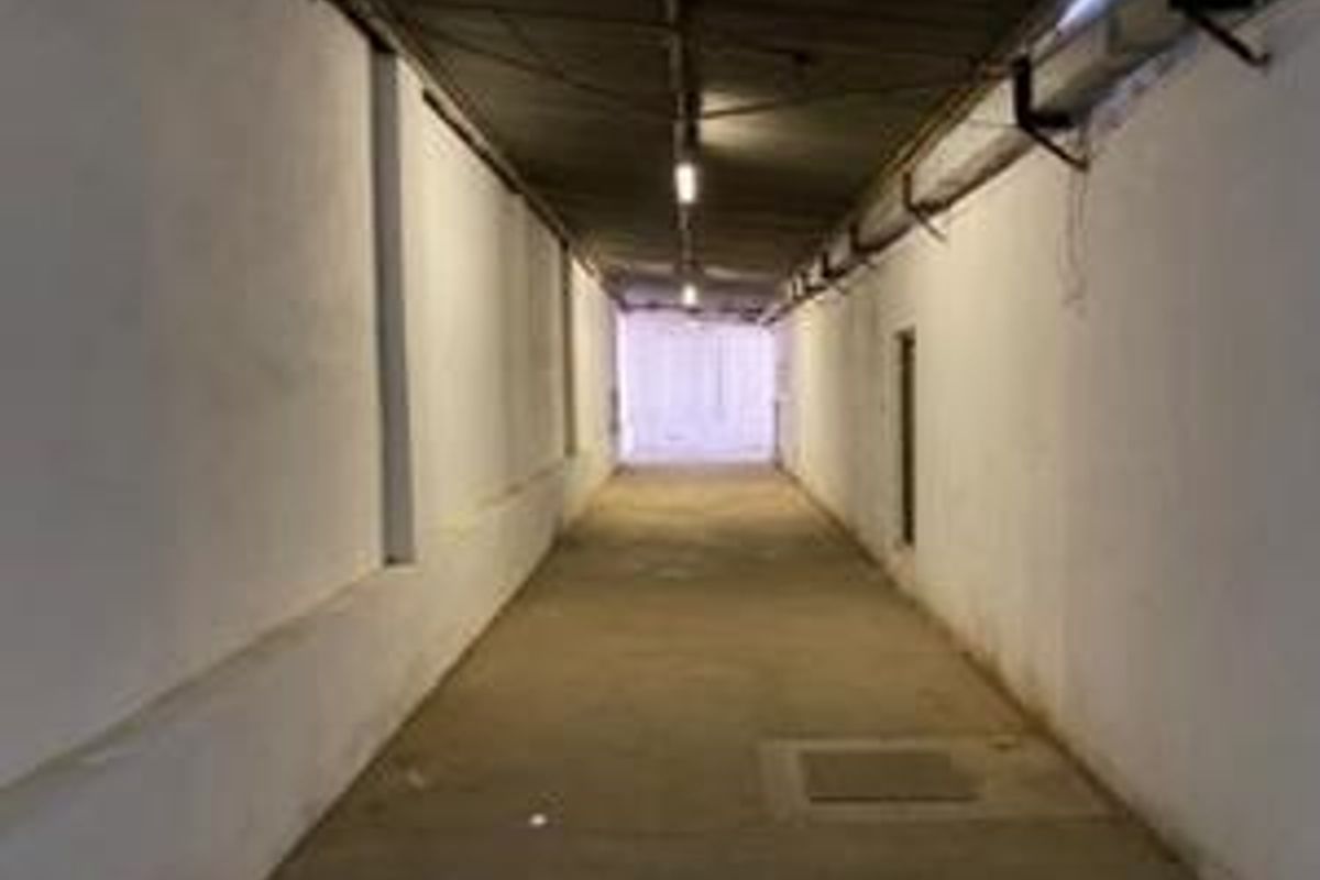 A long, industrial-style corridor featuring exposed ceiling infrastructure, concrete flooring, and basic fluorescent lighting, suitable for various commercial or storage access purposes.