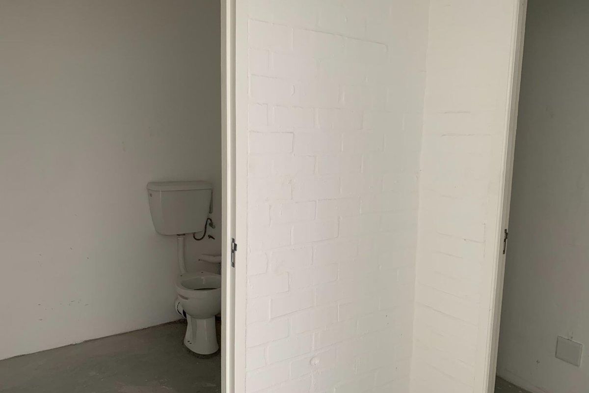 A basic, unfinished bathroom space featuring a standard toilet, white painted brick walls, and an exposed concrete floor.