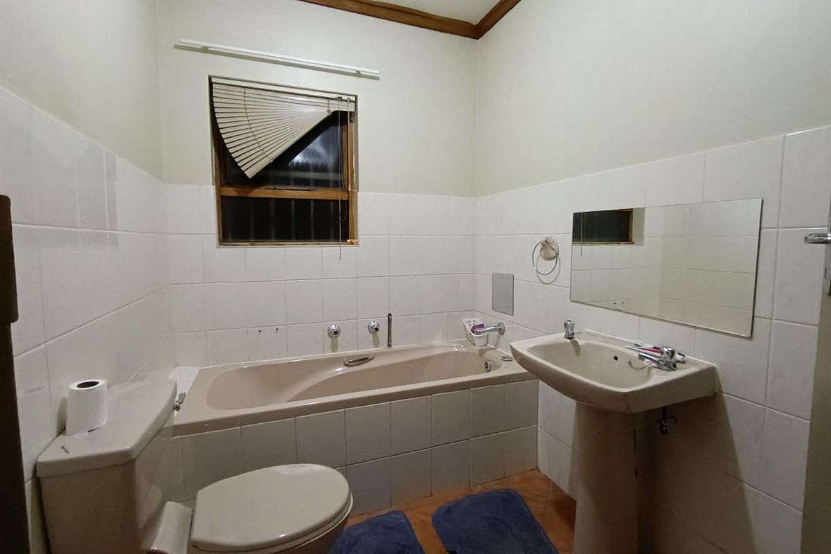 A dated bathroom featuring a beige bathtub, pedestal basin, and toilet, with white wall tiling and a window with a damaged blind.