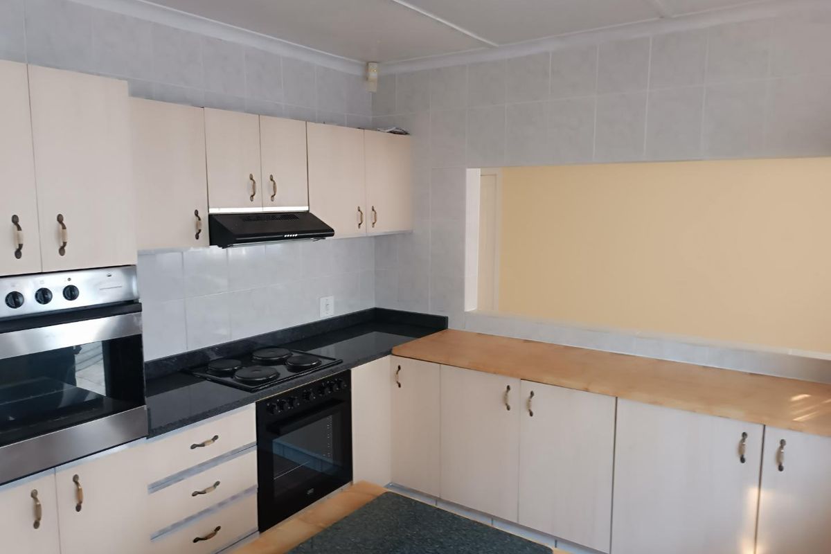 A functional kitchen featuring light-toned cabinetry, dark stone countertops, a built-in oven, electric hob, and an extractor fan with a convenient serving hatch.