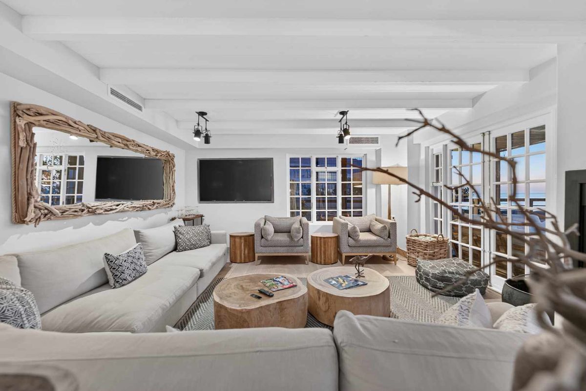 A bright and airy living room featuring exposed white ceiling beams, large multi-pane windows, and direct access to an outdoor area through french doors.