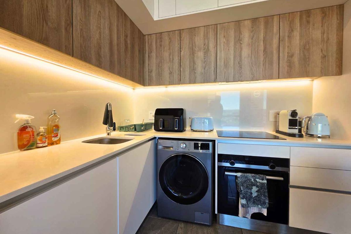 Modern kitchen featuring sleek wood-grain and white cabinetry, stone countertops, integrated appliances, and ambient under-cabinet lighting.
