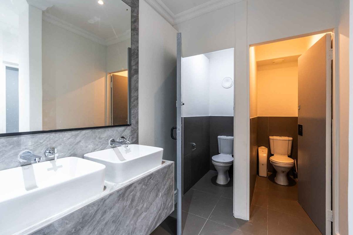 Modern office bathroom featuring a double vanity with vessel sinks, large mirror, and two separate enclosed toilet cubicles with tiled finishes.