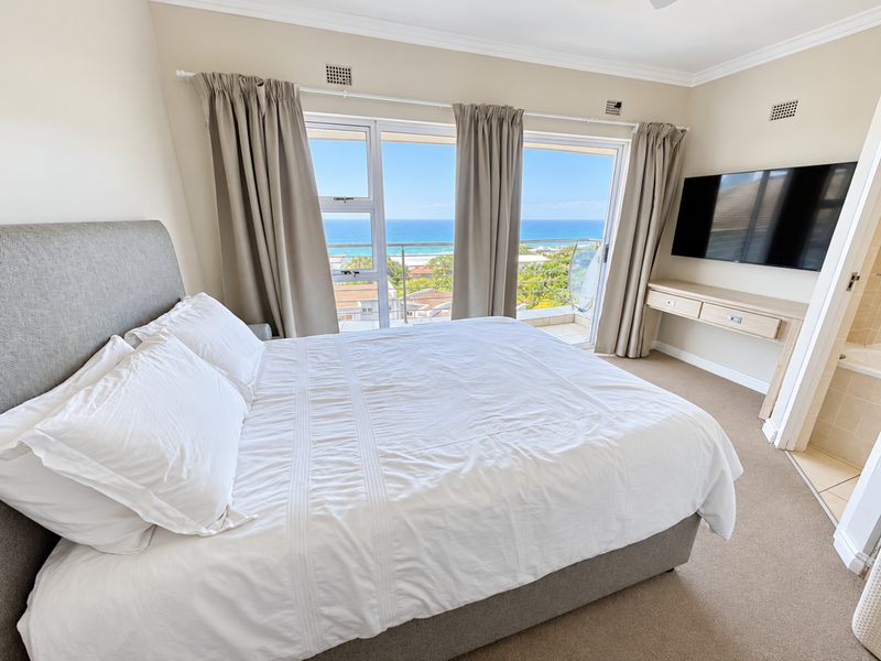 A well-maintained bedroom featuring a built-in vanity, wall-mounted television, and large sliding glass doors that open to a balcony with ocean views.