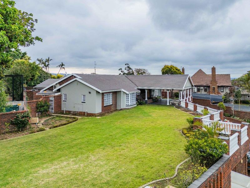 Rear view of the property featuring a spacious lawn, covered patio area, and established brick boundary walls with decorative balustrades.
