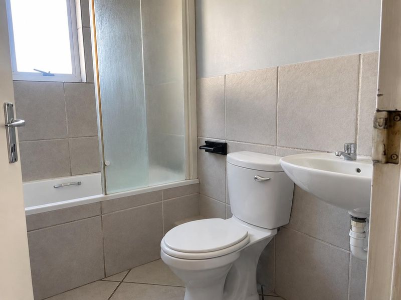 A functional bathroom featuring a bathtub with a shower screen, a standard toilet, and a wall-mounted basin with neutral wall and floor tiling.