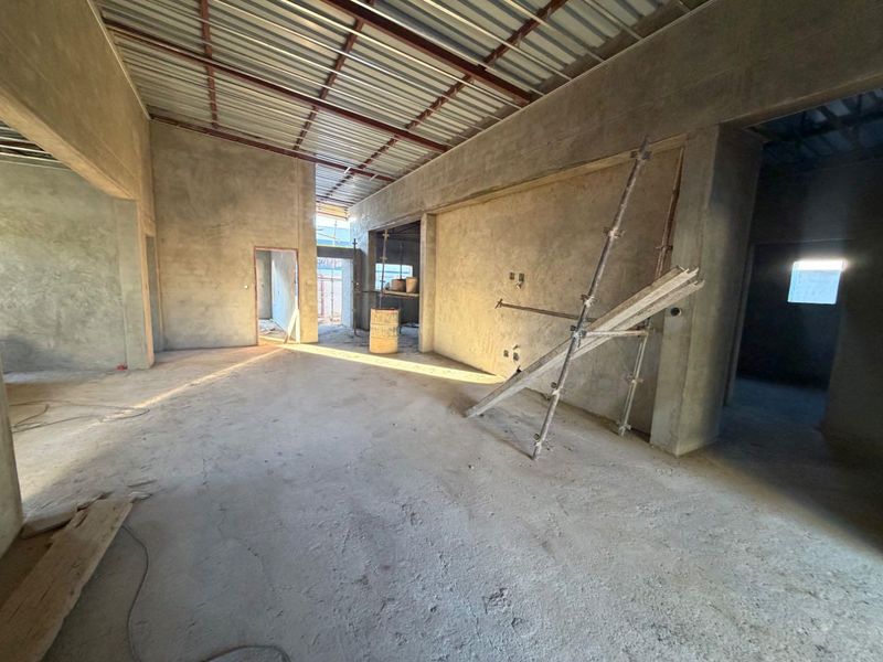 An expansive open-plan interior space currently under construction, featuring exposed metal roofing, plastered walls, and raw cement flooring.